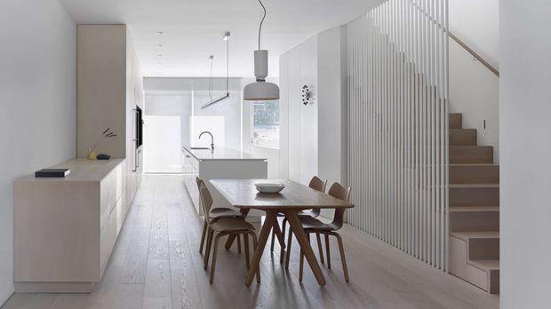 Bright, minimalist open-plan dining and kitchen interior with a light timber dining table and chairs in the foreground, white integrated cabinetry, pale timber flooring, pendant lighting, and a staircase screened by vertical white slats. The space has a clean, airy contemporary appearance with a restrained neutral palette.