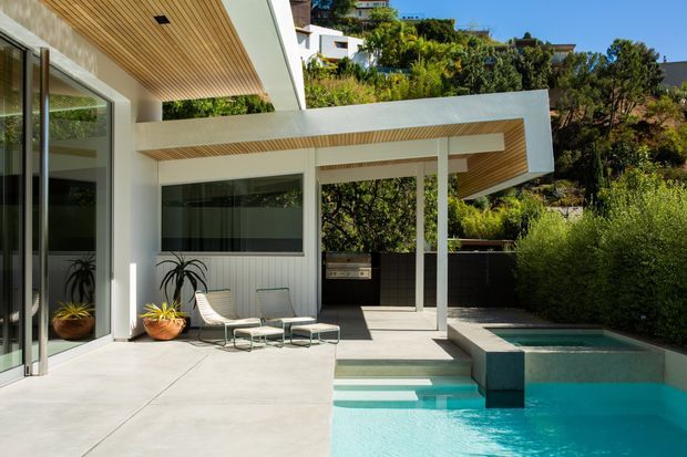 Modernist residential outdoor entertaining area with a turquoise swimming pool beside a white-rendered house. The composition features broad concrete paving, full-height glazing, slender white columns, projecting flat roof forms, and warm timber slatted soffits. A compact covered outdoor area includes lounge seating and a built-in barbecue, with dense landscaped planting surrounding the pool and house.