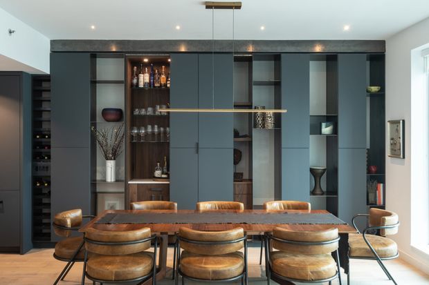 A contemporary dining area dominated by a large dark built-in wall unit with concealed cabinetry, open display shelving, integrated bottle storage, and a central bar display. A long timber dining table with upholstered leather dining chairs sits beneath a slim linear pendant light. The room has pale walls, a light timber floor, dark grey-green joinery, warm wood and leather accents, and abundant daylight from the window at right.