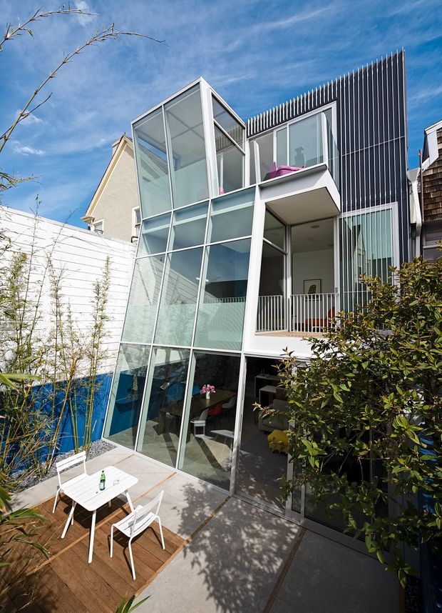 Exterior view of a narrow multi-storey contemporary residence with a dramatic floor-to-ceiling glazed addition, white structural framing, projecting balconies, and a dark vertically ribbed facade. A small timber patio with outdoor dining furniture occupies the foreground, with planted landscaping around the building.