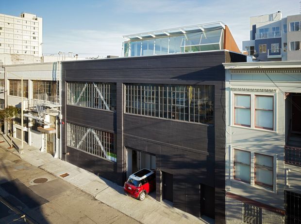 Exterior street view of a contemporary dark-clad multi-storey urban building, featuring large industrial-style barred glazing, a rooftop glazed pavilion or clerestory, and an integrated vehicle entrance. It sits tightly among neighbouring buildings, with an older light-coloured adjoining facade visible at right.