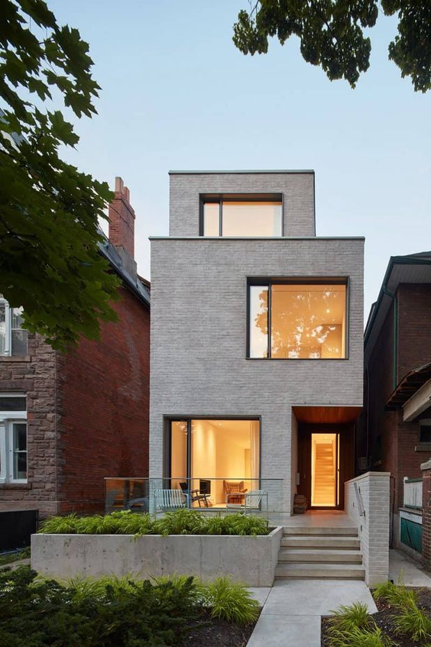 Front exterior view of a narrow, multi-storey urban residence. The contemporary facade is composed of pale grey brick with large dark-framed windows, a recessed timber-lined entry, concrete steps, a glass balustrade, and a landscaped front garden. Warm interior lighting is visible through the glazing, with neighbouring traditional brick homes closely adjoining the property.