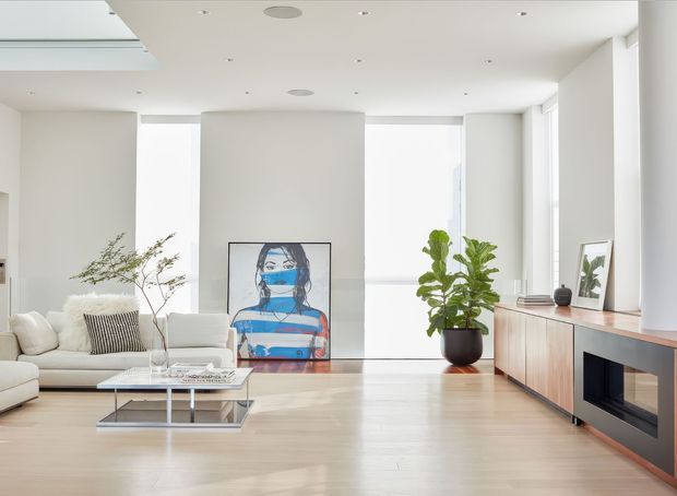 Bright, spacious contemporary living room with a low white sectional sofa, glass-and-metal coffee table, pale timber flooring, large floor-to-ceiling windows, and a long timber media console with a built-in fireplace. White walls and ceiling create an airy setting, complemented by indoor plants and a colourful framed artwork.