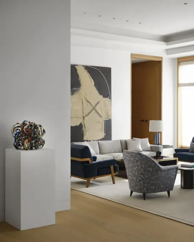 A refined living room with a neutral upholstered sofa, navy and grey armchairs, a low coffee table, and a large pale rug over warm timber flooring. White walls and a layered recessed ceiling are paired with natural timber door and window frames. A large abstract artwork, sculptural object on a pedestal, and table lamp add visual interest.