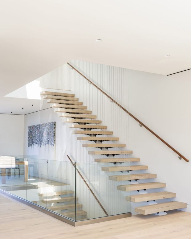 Bright contemporary interior focused on a sculptural floating staircase. Pale timber treads project from a vertically grooved white wall, with slender timber handrails, clear glass balustrades, and a light timber floor. The stair overlooks a lower-level void, creating an airy, open composition with minimal detailing and recessed ceiling lights.