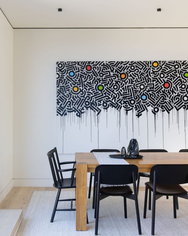 Contemporary dining room with a large rectangular timber dining table surrounded by black chairs. A bold black-and-white abstract artwork with colourful circular accents dominates the rear wall. The room has pale walls, light timber flooring, a light neutral rug, a simple white ceiling with recessed black downlights, and minimal table styling.
