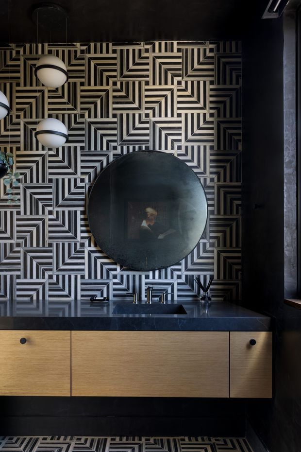 A dark, dramatically styled bathroom vanity featuring a bold geometric black, white and beige patterned tile wall and matching floor. The floating timber vanity has simple black knobs, a dark stone benchtop, integrated basin and black tapware. A large round dark mirror is centered above the vanity, with spherical wall lights and black-painted surrounding walls.