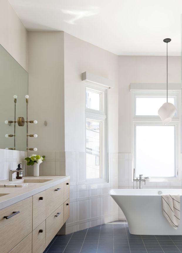 Bright, refined bathroom featuring a freestanding white bathtub beneath tall white-trimmed windows, a large wall mirror, a pale timber double vanity with black hardware, white tiled wainscoting, dark grey floor tiles, and a sculptural pendant light. The space has high ceilings and a clean contemporary-traditional feel.