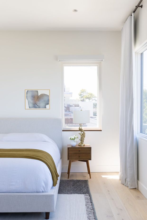 Bright, serene bedroom with a pale upholstered bed, white bedding, and an olive-gold throw. The room has light timber flooring, soft off-white walls, tall sheer curtains, large windows, a compact mid-century-style timber bedside table, and a sculptural lamp. A small abstract artwork adds a subtle accent above the bed.