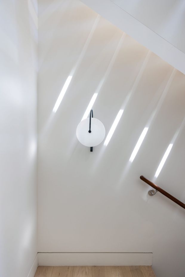 A minimalist stairway or landing with white walls and a sloping paneled ceiling featuring narrow linear skylights or roof lights. A simple round wall clock is mounted centrally, and a dark timber handrail runs along the right wall. The floor is pale timber, creating a bright, restrained interior.