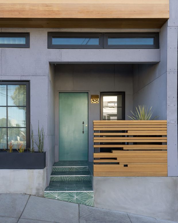 Front entry facade of a compact contemporary residence, featuring a recessed teal front door, narrow clerestory windows, exposed smooth concrete or fibre-cement wall panels, a warm timber overhang, and a horizontal timber privacy screen. Patterned green geometric tiles line the entry steps, with integrated planter boxes and architectural grasses.