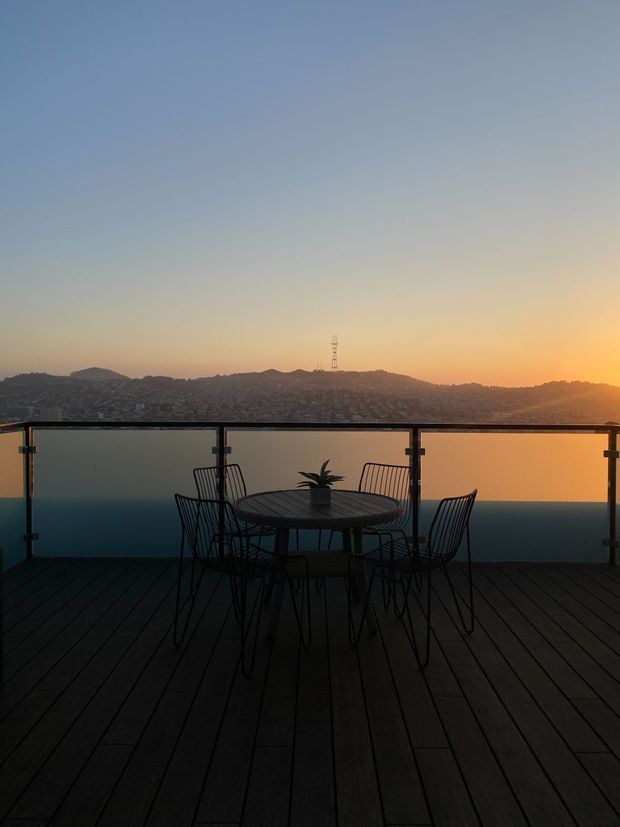 A dark timber-decked balcony or terrace overlooking a hilly urban landscape at sunset. A black metal outdoor dining table with matching wire-frame chairs and a small potted plant sits against a glass balustrade. The scene has strong sunset backlighting, a warm horizon, and a calm, silhouetted atmosphere.