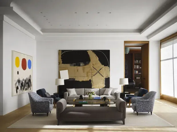 A refined contemporary living room with a symmetrical seating arrangement, including a large sofa, two armchairs, a rear sofa, coffee table, and floor rug. White walls and a recessed tray ceiling with numerous downlights create a bright, polished setting. A large abstract artwork forms the main focal point, while timber-framed glazing and a timber-lined doorway open toward a darker adjoining library or study.
