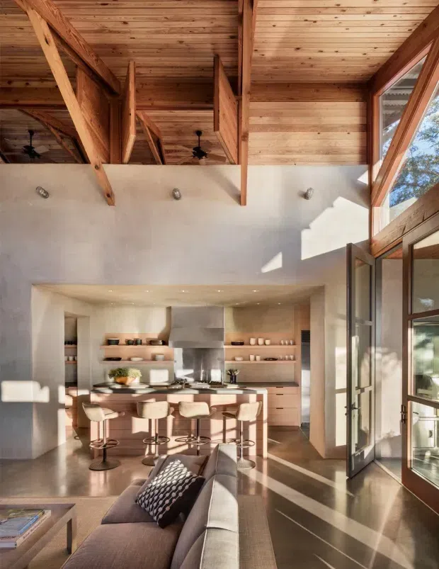 A spacious open-plan interior combining a living room and kitchen beneath a dramatic exposed timber roof structure. The kitchen features a large island with seating, pale wood cabinetry, open shelving, and a stainless-steel rangehood. A contemporary sofa occupies the foreground, while tall glazed doors and high-level glazing bring in abundant natural light. The room has polished concrete flooring, warm timber ceilings and beams, and textured light plaster walls.