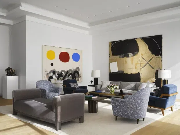 Bright, refined contemporary living room with white walls, substantial decorative ceiling moulding, recessed spotlights, and a large seating arrangement around a dark wood coffee table. The room includes grey upholstered lounge chairs and sofa, patterned blue-grey accent chairs, blue occasional seating, modern table lamps, abstract artwork, a pale area rug, and light timber flooring.