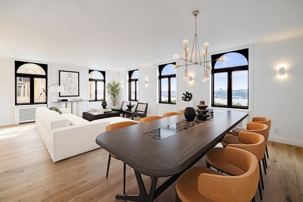 A bright, spacious open-plan living and dining room with tall arched dark-framed windows overlooking a waterfront or urban landscape. The room features pale timber flooring, white walls, a large dark timber dining table with sculptural upholstered dining chairs, a light sectional sofa, minimalist artwork, and a contemporary chandelier. The overall feel is refined, airy, and contemporary with strong architectural window detailing.