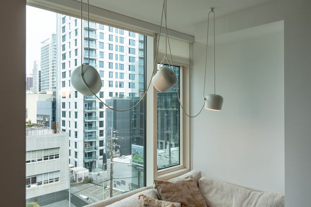 Bright contemporary apartment living area with a large corner of floor-to-ceiling glazing overlooking dense high-rise urban buildings. A pale textured sectional sofa occupies the foreground, while sculptural suspended pendant lights hang at varied heights. White walls and light timber window frames create an airy, minimal interior.