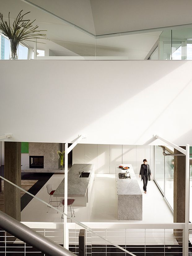 Bright, double-height contemporary interior viewed from an upper stair or landing. The main space is an open-plan white kitchen with two long stone islands, integrated appliances, extensive glazing, and a minimalist dining/living area beyond. White steel or painted structural framing, horizontal metal balustrades, mirrored upper surfaces, and strong natural light emphasize the airy multi-level design.