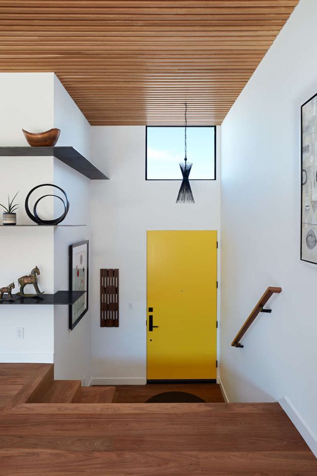 A bright, contemporary residential stairway and entry landing with warm timber flooring and a slatted timber ceiling. White walls frame a bold yellow entry door, black floating display shelves with sculptural decor, a narrow vertical window, pendant light, framed artwork, and a simple timber-and-black handrail descending the stairs.
