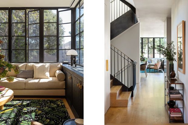 A split-view interior showing a bright contemporary living room and adjoining hallway with a staircase. The living area features large black-framed windows, a cream sofa, dark built-in cabinetry, a sculptural table lamp, and a patterned rug. The hallway has pale timber flooring, white walls, a black metal stair balustrade, timber stair treads, and a console displaying decor and books. A second seating area is visible at the end of the hall.
