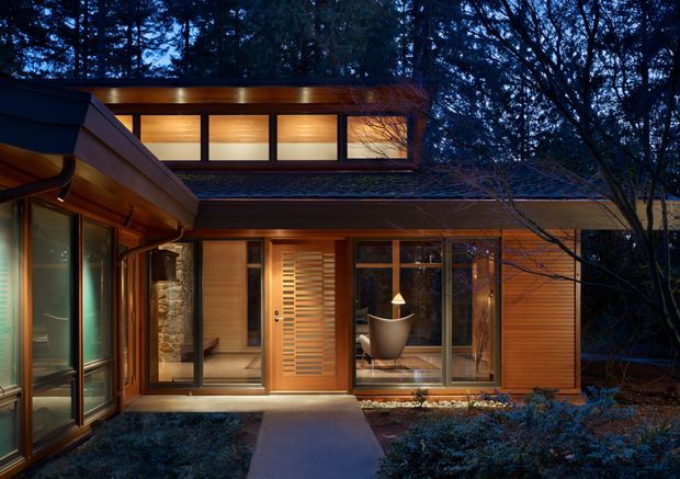 Exterior dusk view of a low-profile timber-clad house nestled among dense trees. The composition shows warm interior lighting through extensive glazing, a covered entry walkway, horizontal timber siding, a glazed projecting wing, and a shallow roofline with clerestory-style upper windows. The scene has a calm, integrated woodland character.