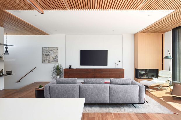 Bright contemporary living room viewed from behind a large foreground surface, with a grey sectional sofa facing a wall-mounted television above a long dark timber media cabinet. The room features warm timber flooring, extensive timber slatted ceiling and soffit detailing, white walls, floor-to-ceiling glazing with curtains, a sculptural lounge chair, and a stair opening at left. Clean-lined joinery and abundant natural light create a refined, warm interior.
