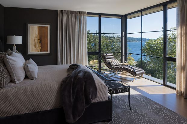 A refined bedroom with a dark feature wall, upholstered bed, layered neutral bedding, bedside lamp, framed artwork, and a lounge chair in a glazed corner sitting area. Floor-to-ceiling windows provide expansive views of trees and a body of water, while patterned carpeting and sheer curtains add softness and texture.