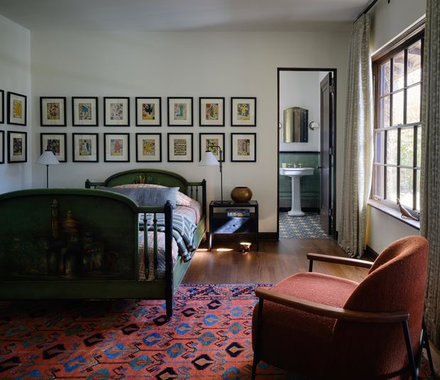 A richly layered bedroom with a dark green upholstered bed, patterned bedding, a gallery wall of framed artwork, timber flooring, and a vivid geometric rug. A rust-coloured lounge chair sits beside the window, while an open doorway reveals a compact bathroom with green wall tile, a pedestal basin, and patterned floor tile. Tall curtained windows bring in natural light.