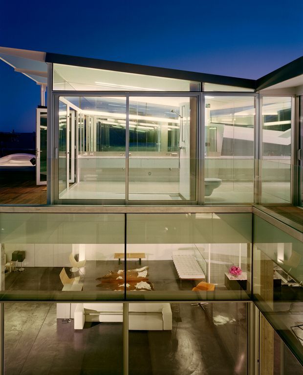 Night-time exterior view of a multi-level contemporary residence dominated by extensive floor-to-ceiling glazing. The upper level contains a bright, glass-enclosed bathroom with visible vanity, mirrors, and toilet, while lower levels reveal a refined living and dining area through the transparent facade. Dark roof planes, slim metal-framed glazing, illuminated interiors, and reflective surfaces create a dramatic modern composition.