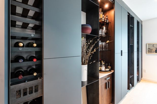 A contemporary built-in storage wall featuring an open wine rack, dark display shelving, and a compact timber-lined home bar with glassware and bottles. Tall matte grey cabinetry and black-framed glazed sections extend along the bright white room, with pale flooring and minimal decor.
