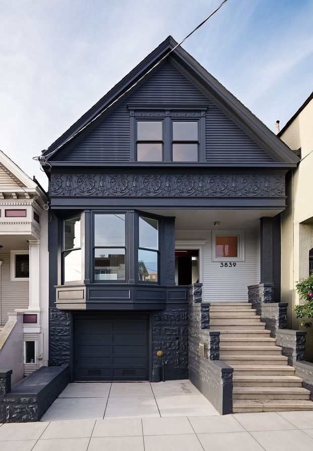 Front elevation of a narrow multi-storey urban house with a steep front gable, dark blue-grey horizontal siding, ornate decorative frieze, projecting enclosed porch, stone-clad lower facade, integrated garage, and a raised stairway to the entry. Dense neighbouring homes frame the facade.