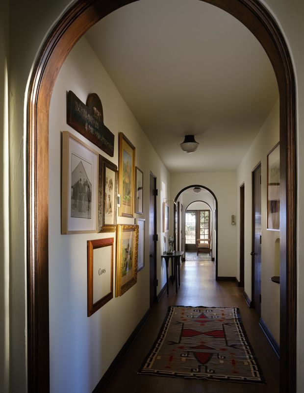 A long, narrow residential hallway viewed through a series of dark-stained timber arched openings. The corridor has pale walls, dark timber flooring and trim, framed artwork along one wall, a patterned runner rug, small console furniture, and traditional ceiling lights. Glazed doors and a bright seating area are visible at the far end, creating depth and natural light.
