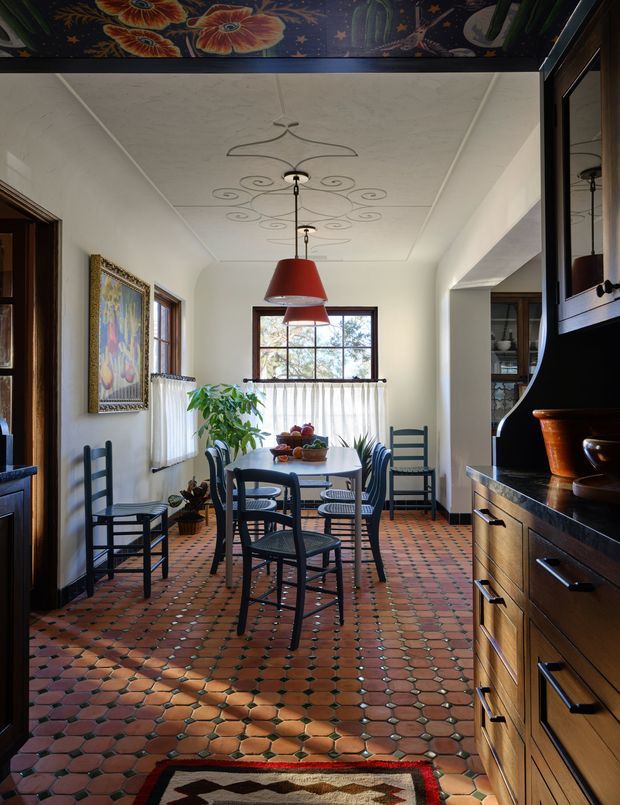 A narrow, characterful dining room with a rectangular dining table and dark ladder-back chairs, illuminated by two red pendant lights. The room features a decorative painted ceiling border, white plaster walls, dark timber-framed windows and doors, a patterned terracotta and green tiled floor, artwork, and substantial dark timber built-in cabinetry along the right wall.