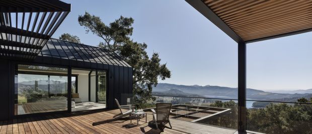 A contemporary residential outdoor terrace with timber decking and a slatted pergola, overlooking a broad lake and mountainous landscape. The terrace connects to a glazed pavilion with dark metal framing and a low, linear roof form. Outdoor seating and a geometric paved area occupy the foreground, while mature trees and the expansive water view create a strong connection to the landscape.