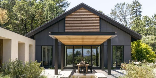 Front exterior view of a contemporary single-storey gable-roofed home. The dark cladding, strong symmetrical facade, exposed timber-lined gable and recessed timber soffit frame expansive glazed doors opening onto a paved outdoor dining area. Established trees and planting surround the building.