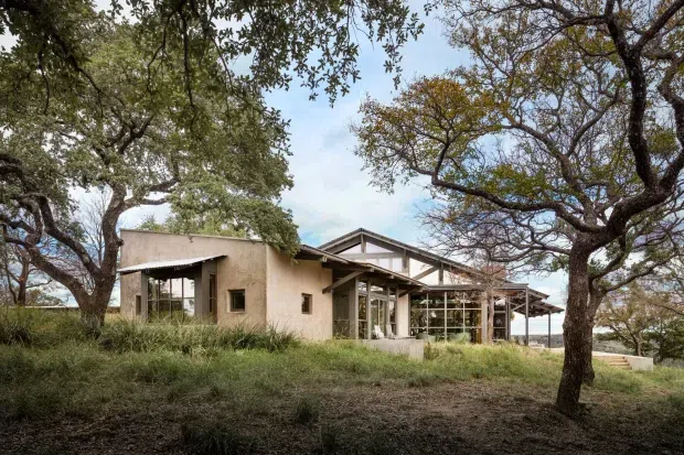 A low, single-storey contemporary house set among mature trees and grasses. The building combines textured beige rendered walls with extensive floor-to-ceiling glazing, exposed timber roof framing, deep roof overhangs, and a glazed pavilion-like living area. The natural, lightly wooded setting and restrained earthy palette give the scene an understated, site-responsive character.