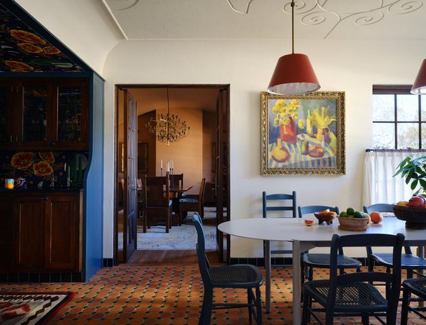 A warm, characterful dining area with a round white dining table and dark woven-seat chairs, opening through timber-framed double doors to a more formal dining room. The room features richly patterned terracotta floor tiles, dark timber built-in cabinetry with decorative painted detailing, ornate plaster ceiling motifs, red pendant lights, framed artwork, and traditional timber windows with sheer curtains.
