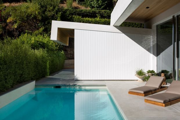 A modern residential pool courtyard with a crisp white sculptural facade, vertical wall cladding, deep overhanging roof forms, and warm timber soffits. Floor-to-ceiling glazing opens onto a pale paved terrace furnished with two timber-framed lounge chairs. Dense clipped greenery borders the turquoise rectangular swimming pool, creating a private, contemporary outdoor setting.