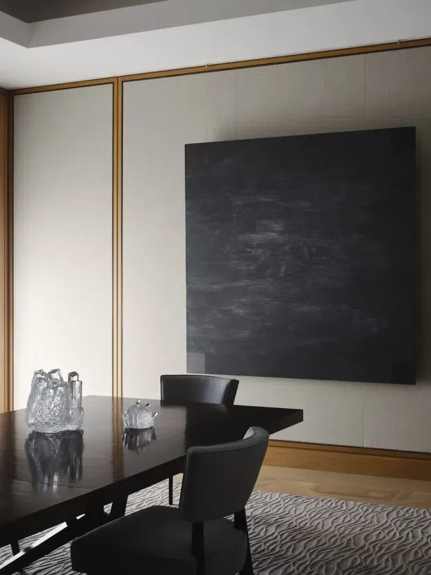 A refined dining room with a large dark rectangular dining table, black upholstered chairs, and sculptural glass or crystal centrepieces. The room features pale vertically textured wall panelling framed with warm timber trim, a dark ceiling accent, timber flooring, and a large monochromatic abstract artwork. A textured grey rug anchors the dining setting.
