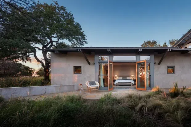Exterior view of a low, single-storey modern dwelling set within landscaped grasses and mature trees. The rendered facade has a broad covered opening with timber-framed glazed doors folded open, revealing a bedroom or living space inside. A shallow corrugated metal roof with exposed dark structural brackets extends over a concrete terrace, with small square windows punctuating the facade.