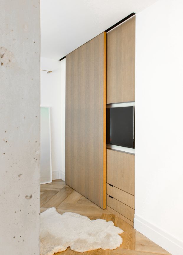 Minimal contemporary interior featuring a full-height light timber built-in partition and concealed storage wall with integrated television, drawers, and a warm orange-toned vertical accent reveal. The cabinetry stands against white walls beside a raw concrete column, with a light timber herringbone floor and pale rug. A doorway and mirror are visible beyond the partition.