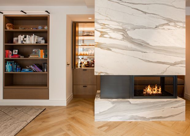 A refined living room featuring a sculptural marble-clad fireplace with an inset gas flame, built-in dark wood shelving, and a concealed bar or display cabinet. The room has warm herringbone timber flooring, white walls, integrated lighting, and a neutral, contemporary palette.