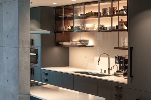 A contemporary kitchen featuring dark matte cabinetry, a pale stone benchtop, integrated appliances, an undermount sink with a curved mixer tap, and open glass-fronted shelving displaying crockery and glassware. Warm under-cabinet lighting, timber accents, and a concrete-look structural wall create a refined, moody atmosphere.
