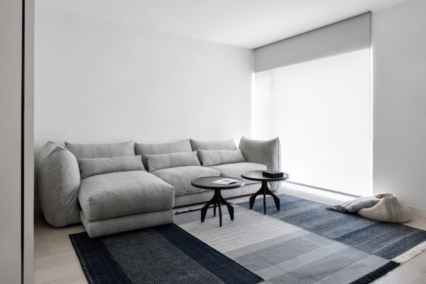A minimalist living room with a large pale-grey sectional sofa, two black round nesting coffee tables, and a broad geometric area rug in layered charcoal, grey, and blue-grey tones. The room has white walls, a white ceiling, light flooring, and a large window fitted with a grey roller blind. A knitted floor cushion rests near the window. The space is bright, restrained, and contemporary.
