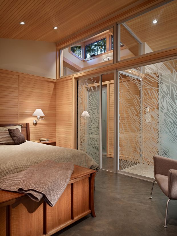 Warm contemporary bedroom with a timber-paneled feature wall and matching slatted timber ceiling. The room includes a built-in timber bed surround, neutral bedding, wall-mounted reading lights, a dark polished concrete floor, and a glazed partition with a decorative etched pattern separating the bedroom from an adjacent space. High-level glazing and clerestory windows bring in natural light, creating an open, refined interior.