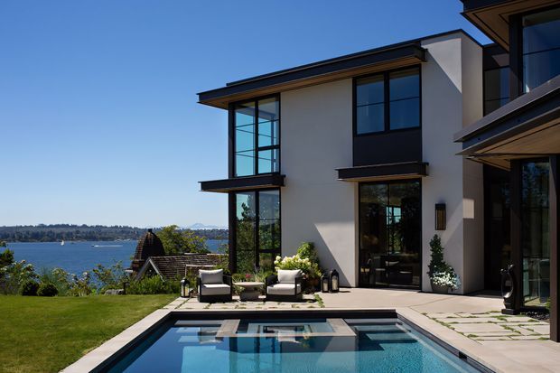 Exterior view of a contemporary multi-storey waterfront residence with clean white and dark trim, extensive floor-to-ceiling glazing, and projecting roof eaves. A landscaped lawn, paved outdoor entertaining area, and rectangular swimming pool occupy the foreground, with a broad body of water visible beyond the property.