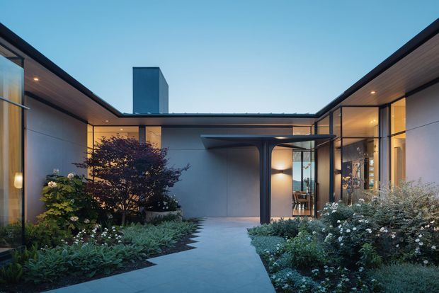 A contemporary single-storey courtyard-style home viewed from the landscaped entry garden. Flat-roofed wings with dark metal framing, expansive glazing, warm timber soffits, smooth light walls, and a sheltered entrance surround a paved pedestrian path and dense planting.