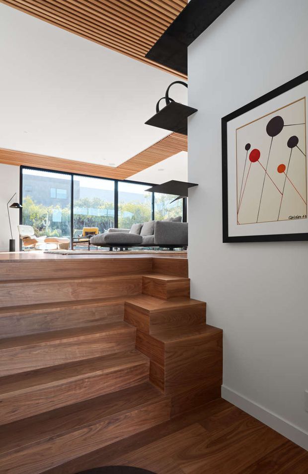 Interior view of a contemporary split-level living area with broad walnut-toned timber stairs and flooring, a stepped transition into a sunlit lounge, full-height black-framed glazing, and a slatted timber ceiling detail. Minimal black pendant lights and a framed abstract artwork complete the refined modern composition.