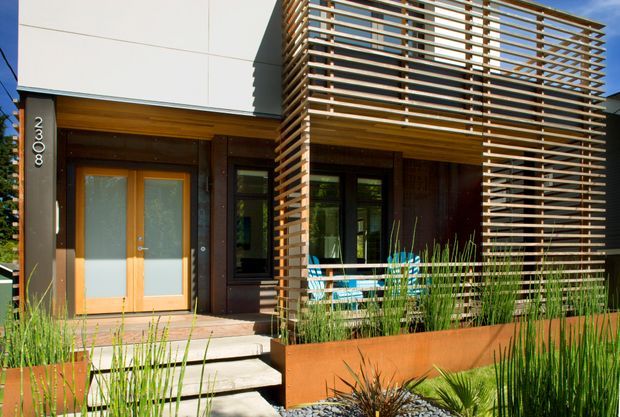 Contemporary residential facade with a light-coloured rectangular upper volume, warm timber entry door and soffit, dark vertical wall cladding, and a prominent horizontal timber-louver screen enclosing a front porch or balcony. A raised corten-like planter with ornamental grasses and drought-tolerant planting borders the entry steps.