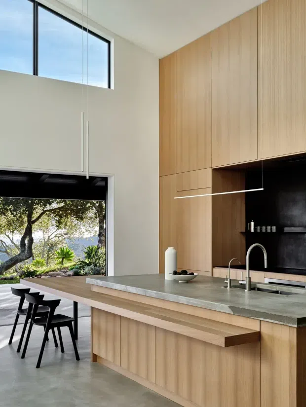 A bright, contemporary kitchen with a large timber-clad wall of integrated cabinetry, a pale stone island bench with timber overhang, black splashback, and minimal black bar stools. Floor-to-ceiling glazing opens to a leafy garden and distant landscape, while a high window brings in additional natural light. The space has clean lines, restrained detailing, and a warm natural-material palette.